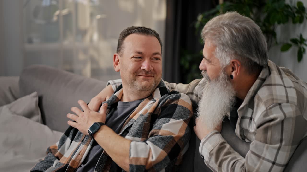 Happy elderly man with gray hair and a lush beard communicates and hugs his brunette boyfriend with stubble in a plaid shirt on a gray sofa in a modern apartment
