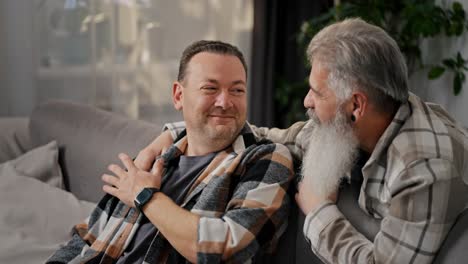 Un-Anciano-Feliz-Con-Cabello-Gris-Y-Una-Barba-Exuberante-Se-Comunica-Y-Abraza-A-Su-Novio-Moreno-Con-Barba-Incipiente-Con-Una-Camisa-A-Cuadros-En-Un-Sofá-Gris-En-Un-Apartamento-Moderno