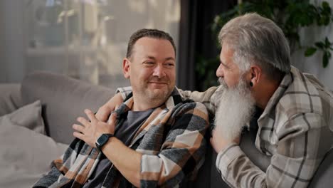 happy elderly man with gray hair and a lush beard communicates and hugs his brunette boyfriend with stubble in a plaid shirt on a gray sofa in a modern apartment