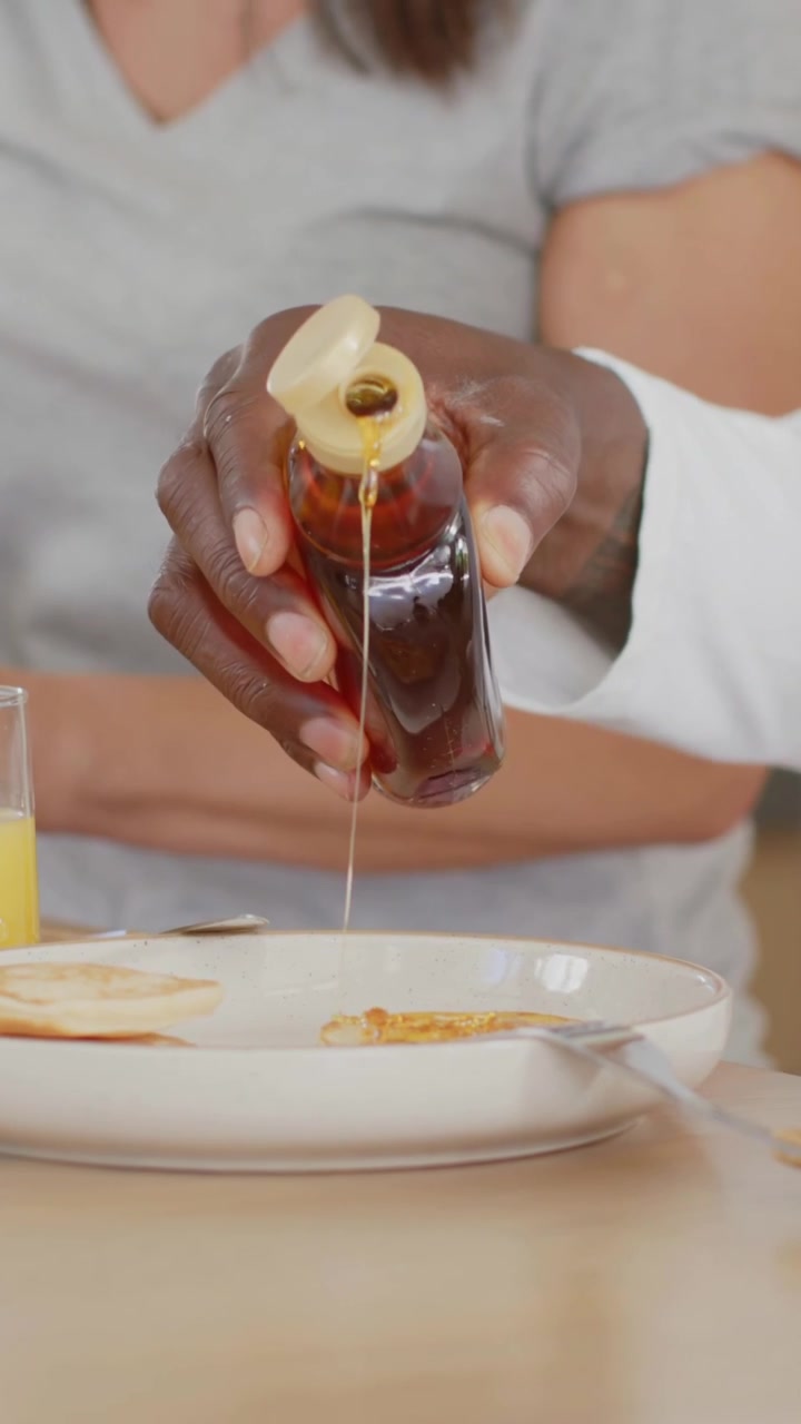 Uomo che versa sciroppo inclinando la bottiglia per irrorare i pancake su un tavolo di legno per colazione, mentre il partner osserva.