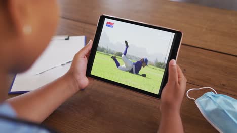 compuesto de una mujer sentada en una mesa con una máscara facial, viendo un juego de béisbol en la tableta