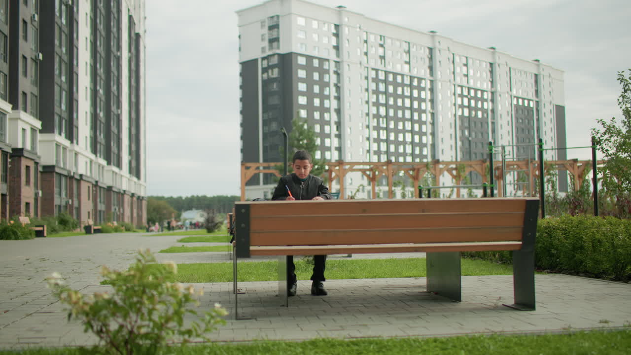 Little boy sits on wooden bench in modern park writing in notebook with focused expression, backpack beside him, tall residential buildings in background under cloudy sky