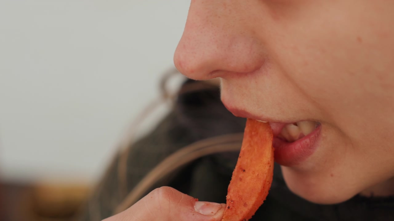 Tampilan close-up wajah bagian bawah dari hidung hingga leher, menangkap aksi makan dengan bibir terbuka, lidah dan gigi bertemu camilan goreng berbumbu, tangan membawa gigitan renyah ke mulut dalam cahaya lembut yang menyebar.
