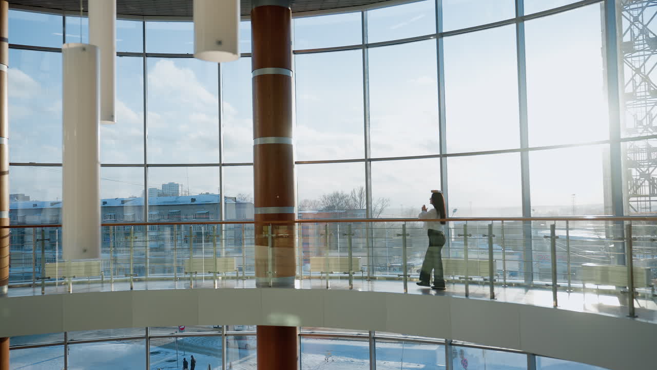 Wide view of stylish modern mall interior with large glass panels and natural daylight as elegant woman walks gracefully across upper floor walkway balancing book on head with confident posture