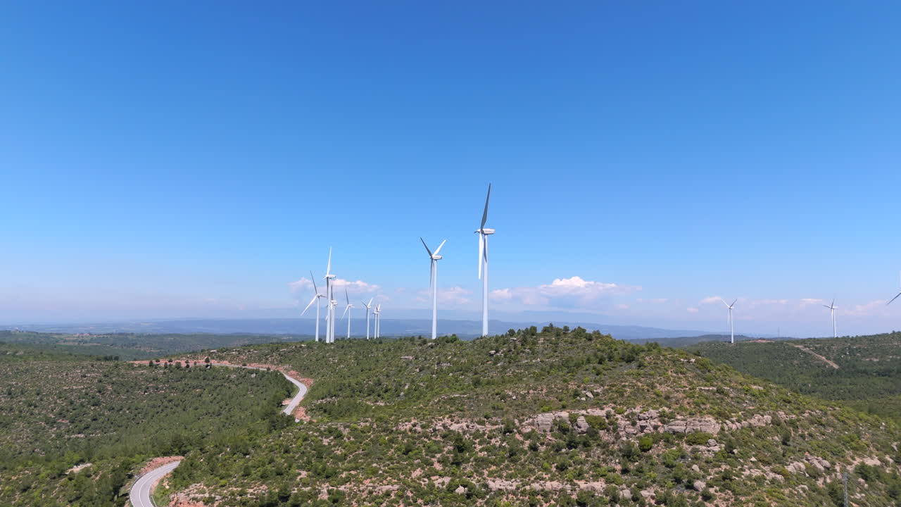 Aerial clip flying forward toward multiple wind turbines spinning on a mountain ridge, surrounded by forest and rocky terrain under a bright blue sky