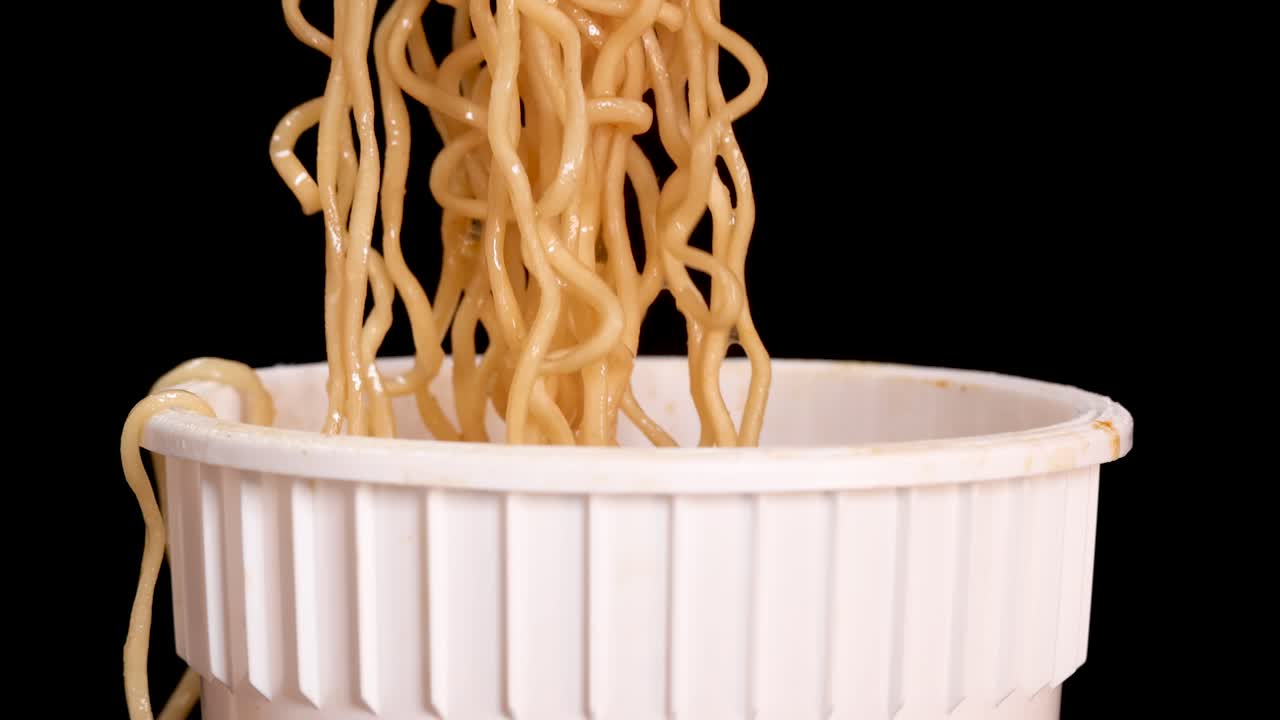 Una mano usa una forchetta per sollevare noodles istantanei cotti da una tazza bianca usa e getta su uno sfondo nero, con un'illuminazione da studio uniforme e una telecamera statica.