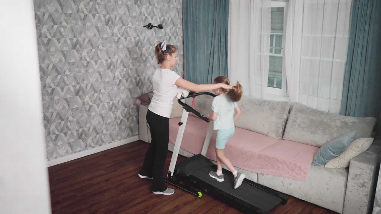 Woman gently wipes sweat from young girl's face using towel while she walks on treadmill near sofa in cozy living room with natural daylight and patterned walls during intense home workout session