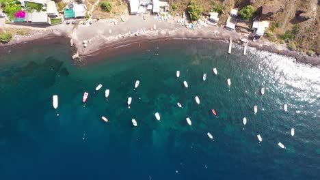 Vista-De-Arriba-Hacia-Abajo-De-Muchos-Barcos-De-Pesca-En-El-Mar-Mediterráneo-Con-Agua-Azul-Cristalina-Y-Playa-Que-Se-Eleva-Alto-Revelando-En-Santorini,-Grecia