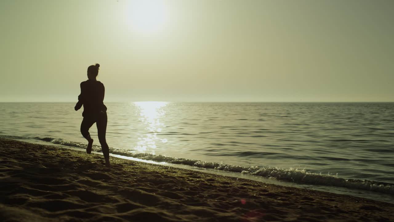 해가 지는 모래 해변에서 혼자 달리는 빠른 여자. 바다 근처에서 조 ⁇ 하는 실루 ⁇  소녀