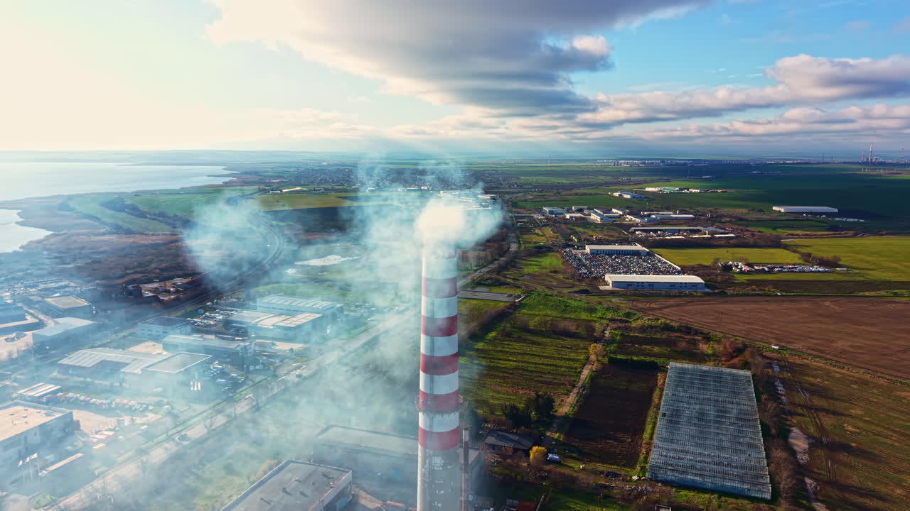 Pollution rises from a factory chimney near a landscape of fields and warehouses during the day with clouds in the sky