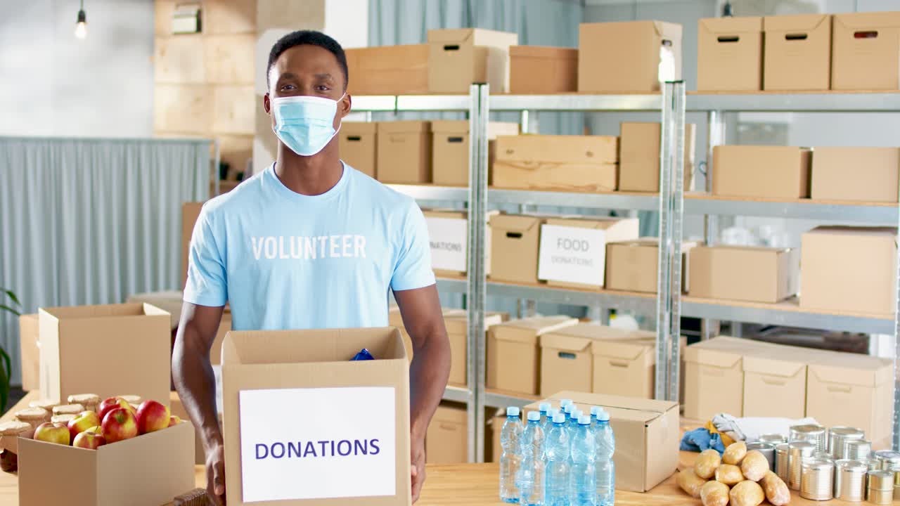 African american male volunteer wearing facial mask holding donation box and smiling to the camera in charity warehouse