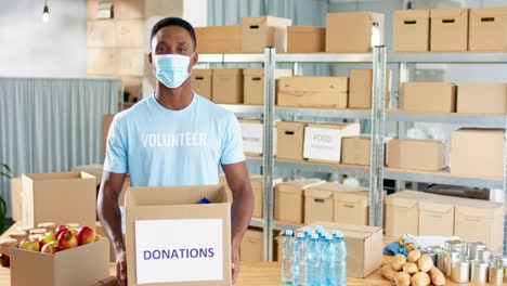 voluntario afroamericano con máscara facial sosteniendo una caja de donación y sonriendo a la cámara en un almacén de caridad