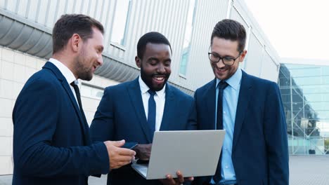 three caucasian and african american businesmen in stylish clothes reading something on the laptop and talking in the street