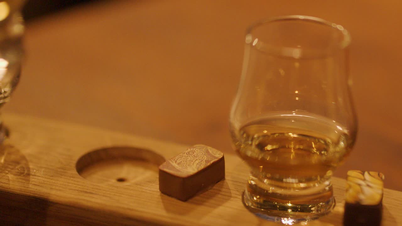 A hand sets a whiskey glass on a wooden tasting board beside chocolate pieces in a warmly lit indoor setting, captured in a close-up sequence