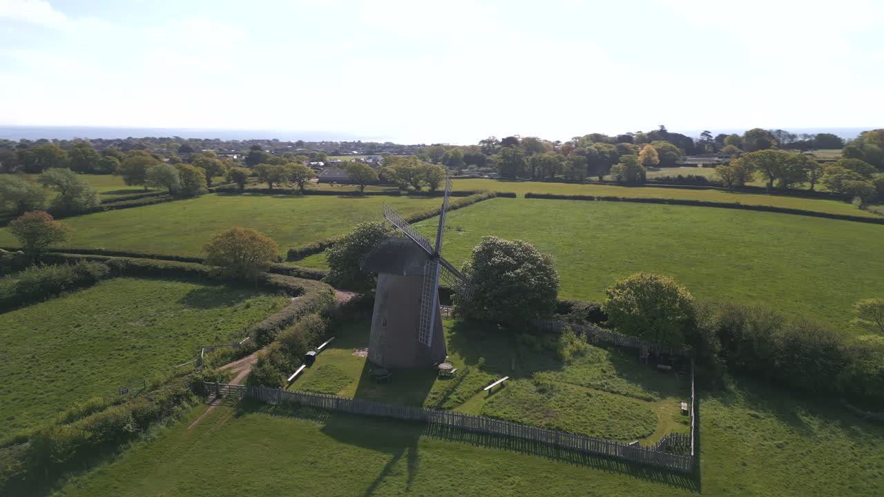 Scenic spinning aerial shot of Bembridge Windmill surrounded by lush green fields, trees, and distant coastal views on a sunny day, Isle of Wight.