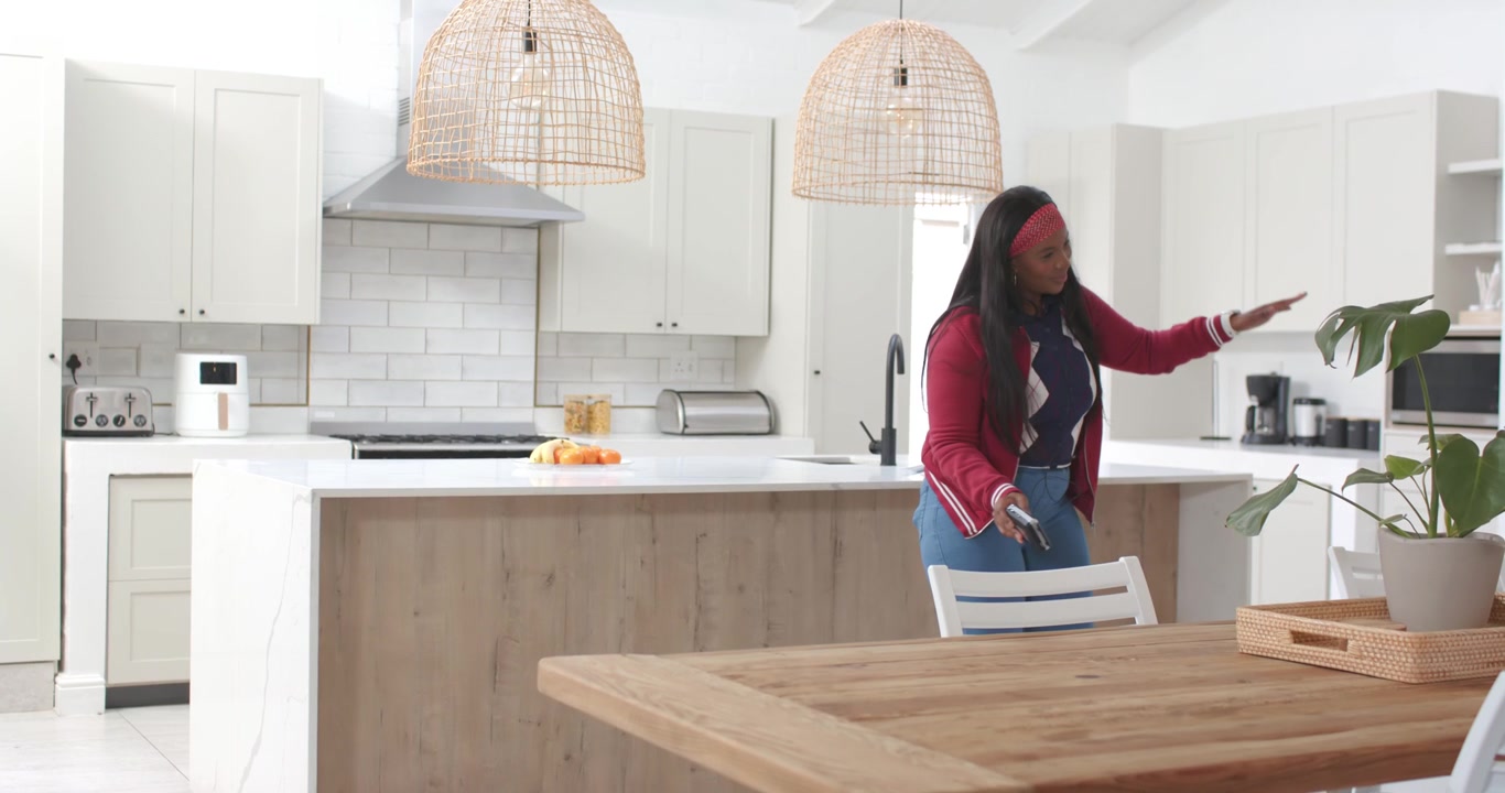 Donna afroamericana che cammina in cucina, alzando il telefono e sollevando una tazza, indicando una pianta, con spazio per il testo