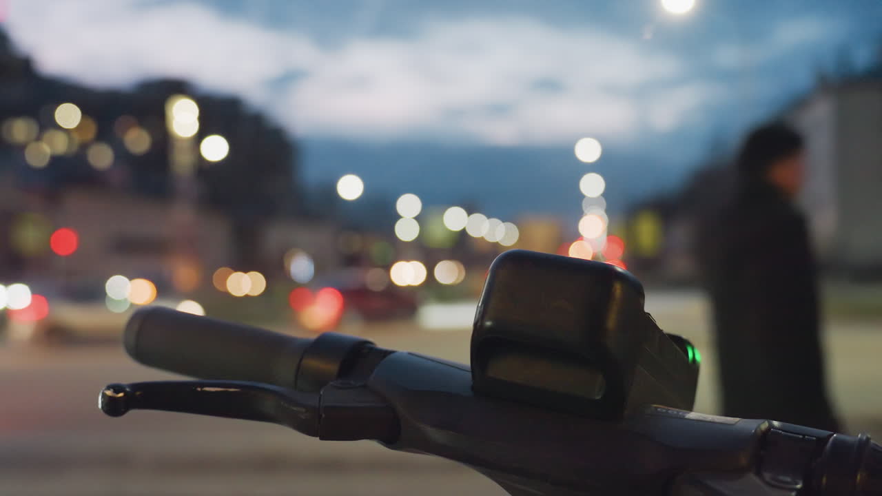 Close up of power bike handle in sharp focus against backdrop of night street glowing with blurry car lights, people passing, and streetlamps illuminating urban road with soft ambient motion