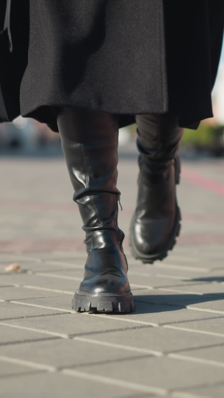 vista de cerca de la pierna de un individuo con botas y abrigo negros, sosteniendo un maletín negro, caminando por un camino entrelazado en un entorno urbano