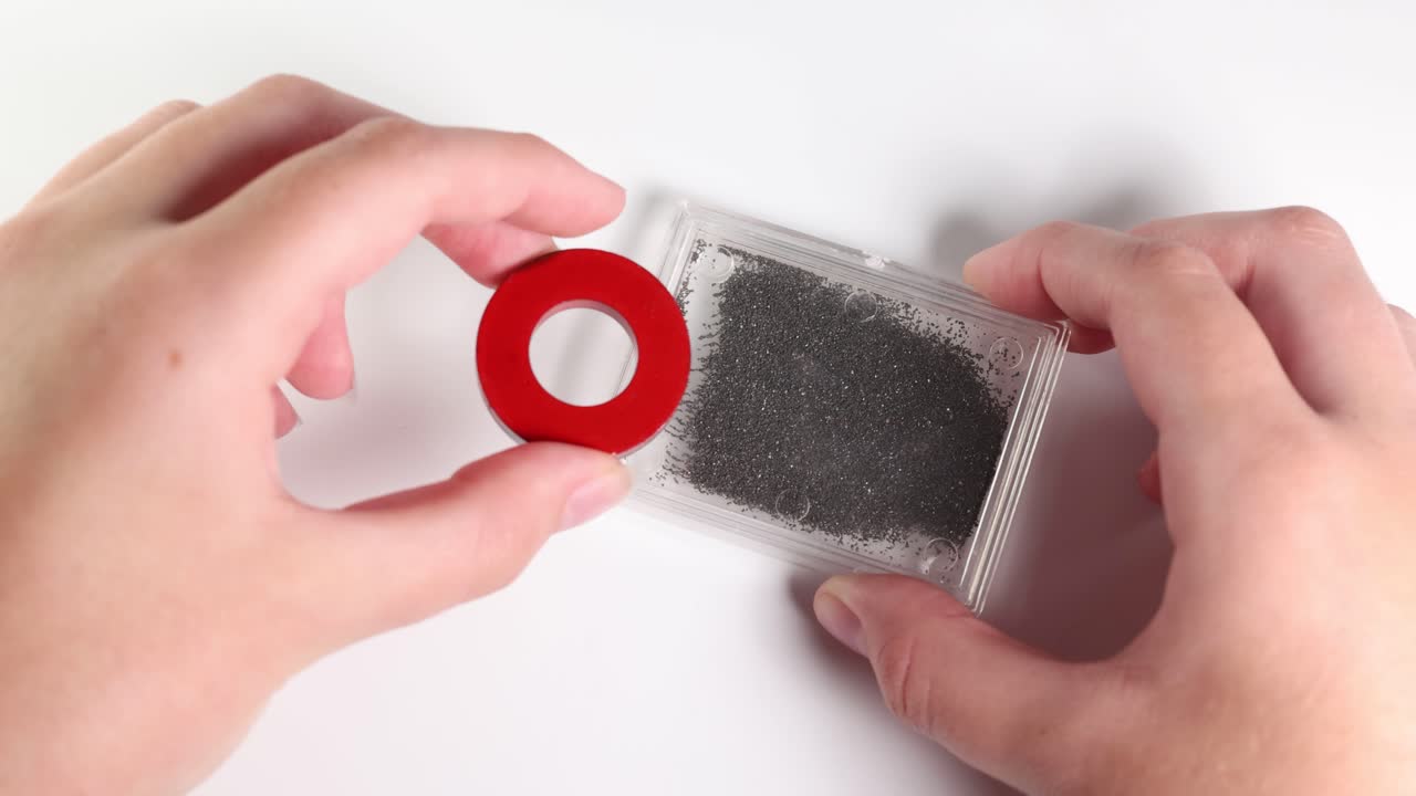 Hands manipulate a magnet over iron filings, demonstrating magnetic attraction and repulsion in a clear, well-lit environment