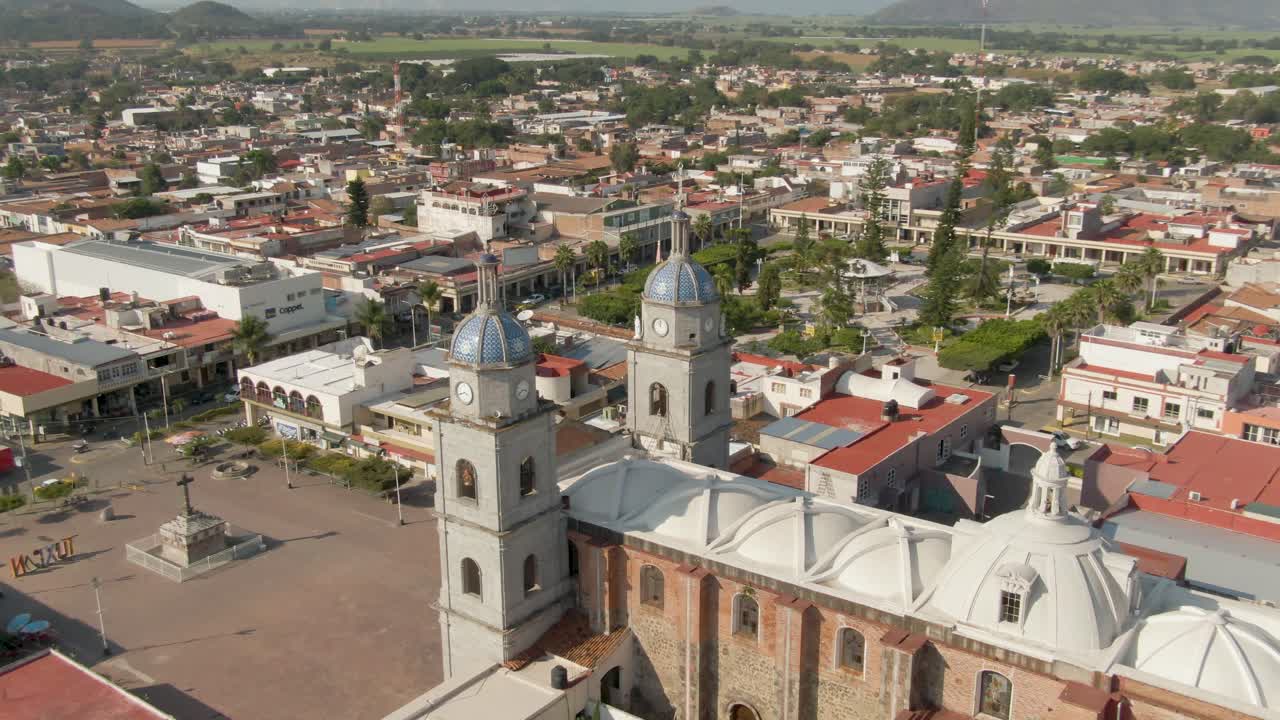 Aerial shot approaching San Juan Bautista Temple roof, orbiting to show central plaza and surrounding buildings in Tuxpan, Jalisco
