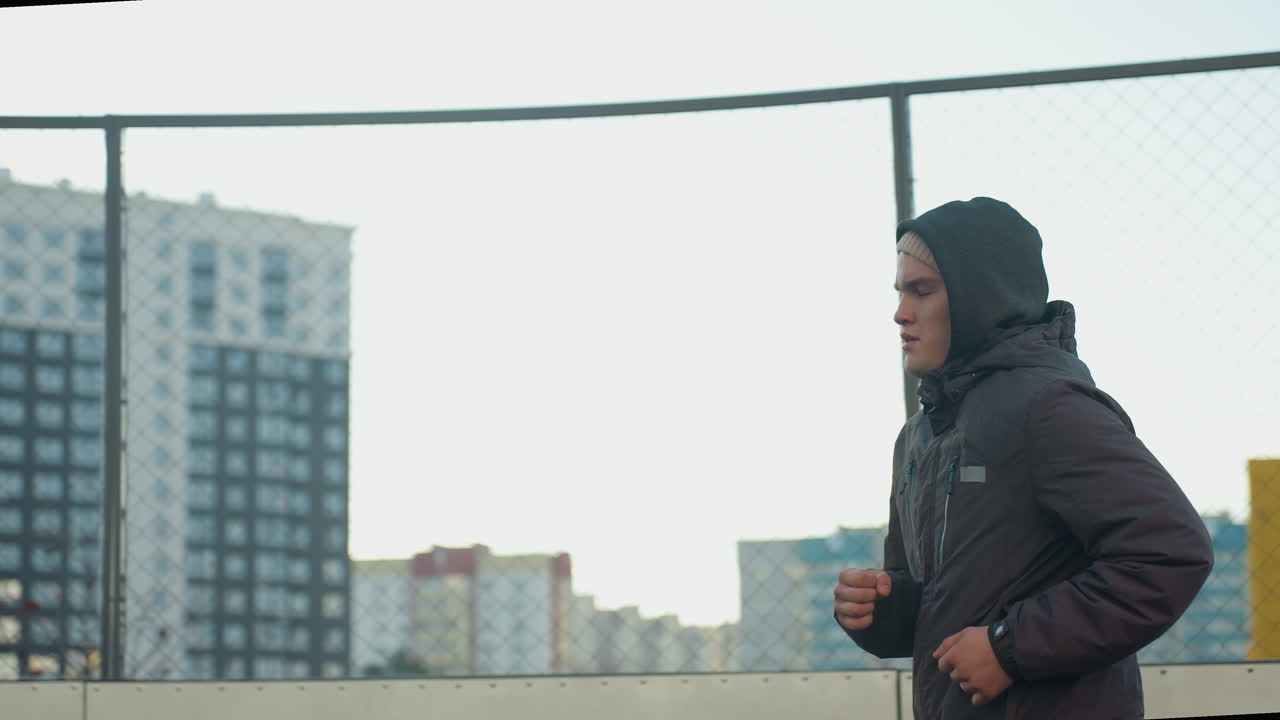 Side view of athlete jogging on outdoor sport arena with focused expression, highlighting determination, fitness, and perseverance against urban backdrop and wire mesh