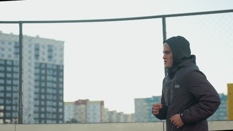 side view of athlete jogging on outdoor sport arena with focused expression, highlighting determination, fitness, and perseverance against urban backdrop and wire mesh