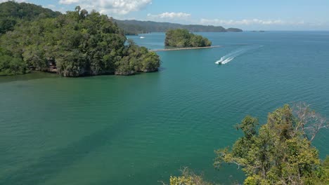 White-speed-boat-drives-on-tropical-ocean-near-jungle-forest,-aerial-orbit