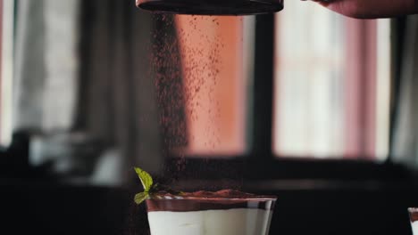 Chef-pouring-cacao-powder-over-a-dessert-dish-of-white-cream-or-yogurt-served-in-a-glass-with-a-mint-garnish