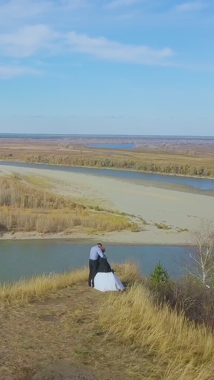 newly wedded couple hugs on steep bank with dry grass near large tranquil river on autumn day upper view