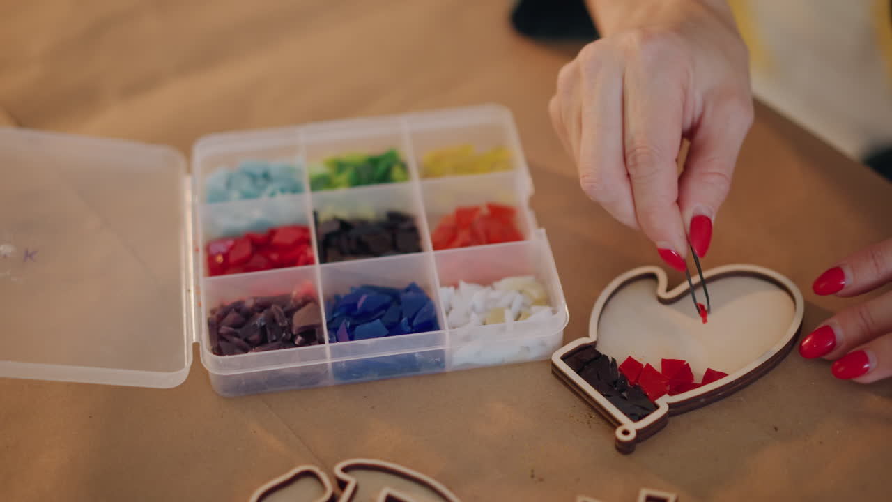 Kvinde fylder hjerteformet ornament med perler, nærbillede af rødlakerede hænder, perleorganizer med flerfarvede rum, præcis placering, detalje til personlig gave, blød sæsonbelysning.