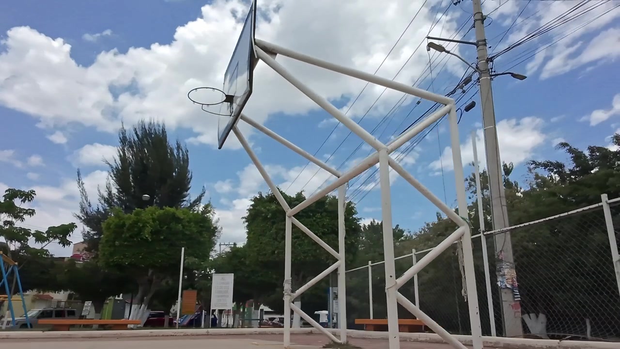 Outdoor basketball court with empty hoop structure standing on paved surface, surrounded by trees and low buildings under a partly cloudy sky in Huatulco Oaxaca Mexico