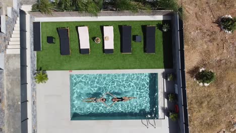 Slim-women-and-men-float-in-a-beautiful-outdoor-pool---she-wears-a-dark-swimming-costume-and-enjoys-the-sun---drone-films-her-from-a-bird's-eye-view