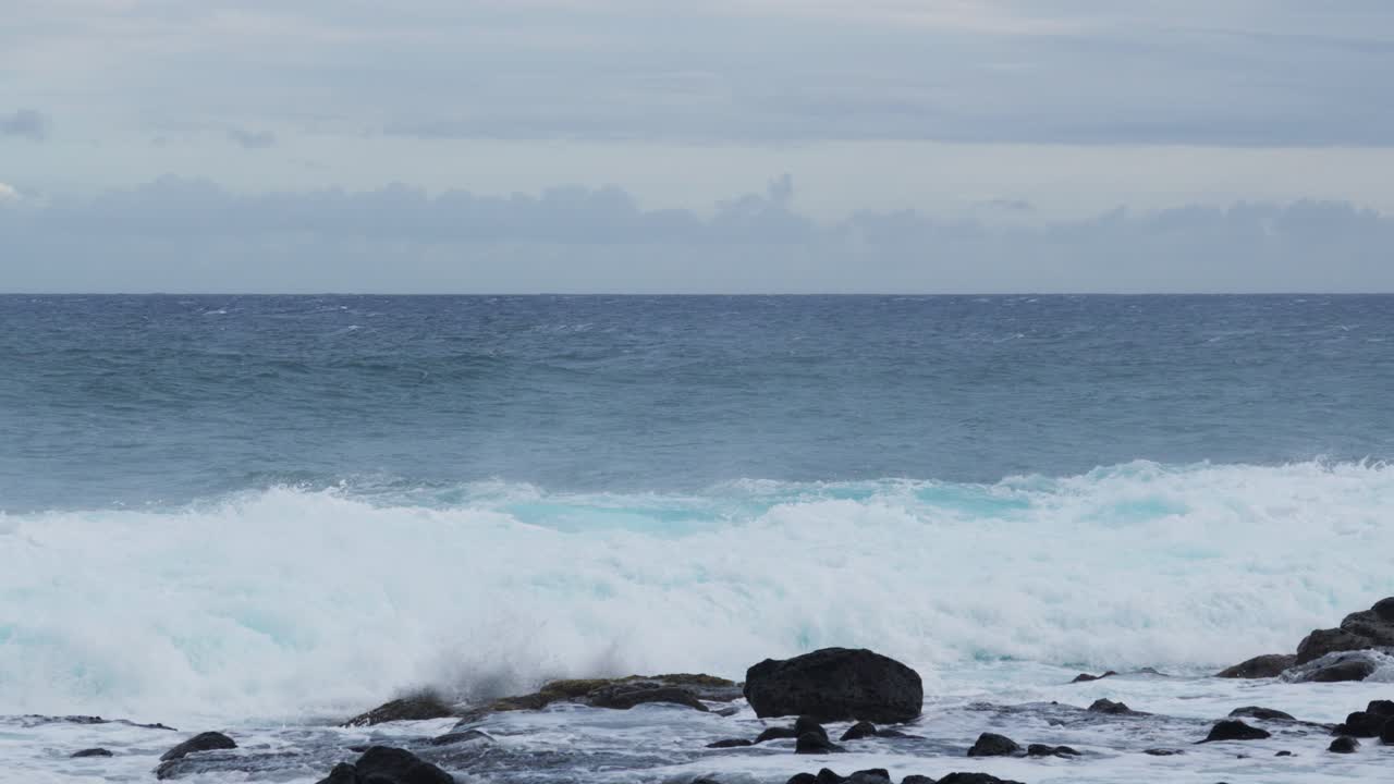 Wind-driven swell detonates against dark shoreline rocks, throwing plumes of white spray as foamy water floods the frame, a moody, restless seascape of raw coastal energy