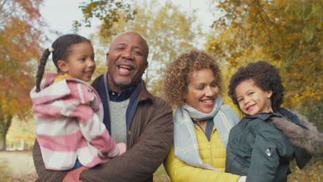 Portrait-Of-Smiling-Grandparents-Carrying-Grandchildren-On-Walk-Along-Track-In-Autumn-Countryside