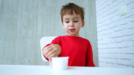 cute toddler in red t-shirt picks a spoon from the table. lovely baby starts to eat yogurt from the cup. low angle view.