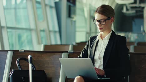 Stilvolle-Und-Erfolgreiche-Geschäftsfrau-Mit-Brille,-Die-Am-Laptop-Arbeitet,-Während-Sie-Auf-Ihren-Flug-Am-Flughafen-Wartet