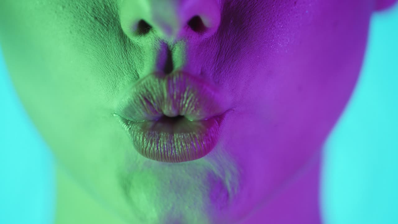 Extreme close up of a beautiful woman lip while she symbolizes a kiss into the camera with green purple background in slow motion