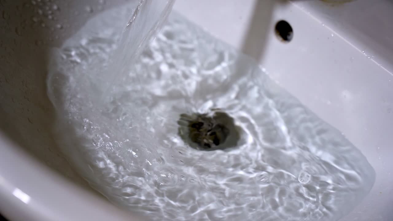 Close-up of clear tap water flowing rapidly into a white ceramic washbasin. Water splashes against the drain, creating ripples, bubbles, and turbulence in a clean bathroom sink