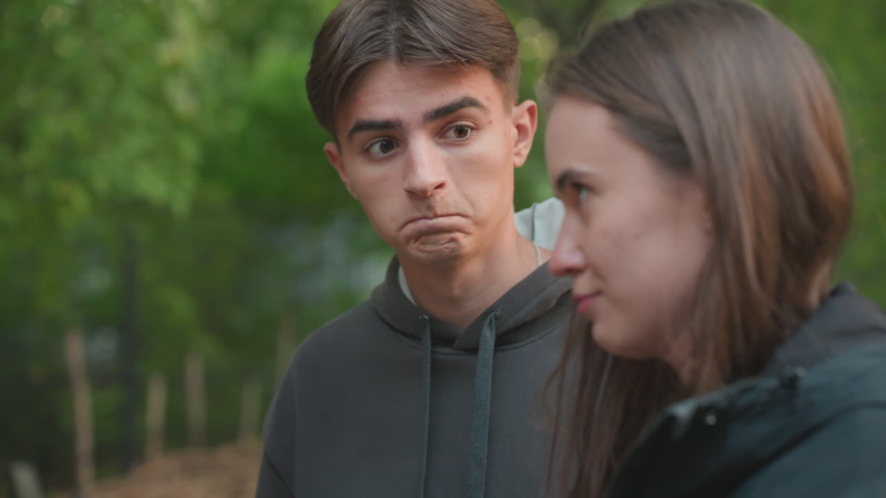 white teenage male listening to friend in park, hoodie and jacket, female closeup with sad expression, empathetic glances and subtle playful reaction from male, candid conversation, natural light