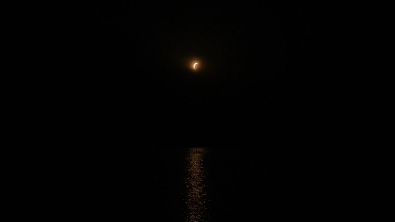 Piccola luna crescente o eclissi parziale brilla sopra acqua nera calma, proiettando un sottile riflesso dorato attraverso l'oscurità, con dettagli di notte, luna, eclissi, falce, che mostrano oscurità, cielo, acqua