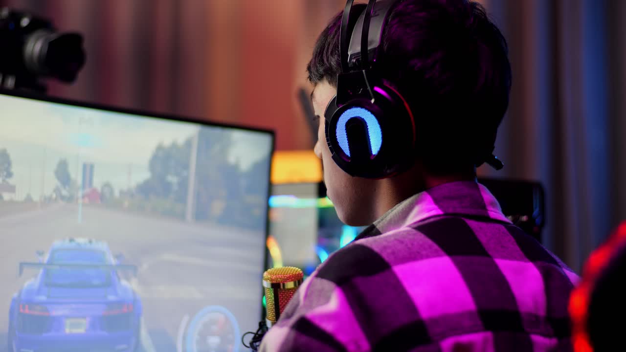 Close Up Back View Of Asian Boy Streamer Playing Car Racing Game Over Network On Computer, Talking On The Microphone.  Live Stream Video Game, Desk Illuminated By Rgb Led Strip Light