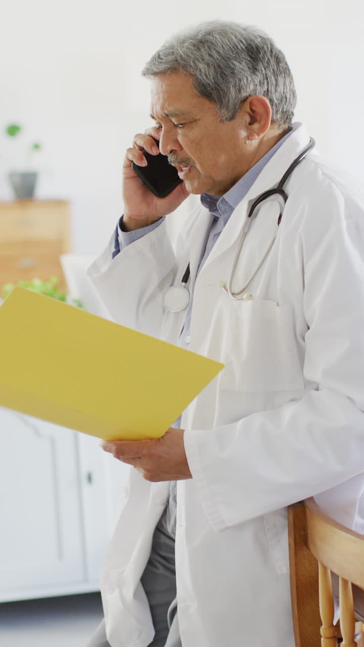 Video verticale di un medico anziano birazziale che tiene in mano una cartella e parla al telefono