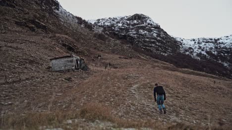 young man hiking through abandoned meadow with small wooden shed and snow-covered mountains in the background