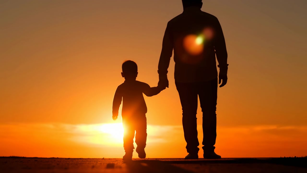 Siluetas oscuras de un adulto y un niño caminan de la mano por un terreno abierto al atardecer, con el niño dando un paso adelante junto al adulto bajo un cielo naranja cálido, escena familiar tranquila desde un punto de vista bajo.