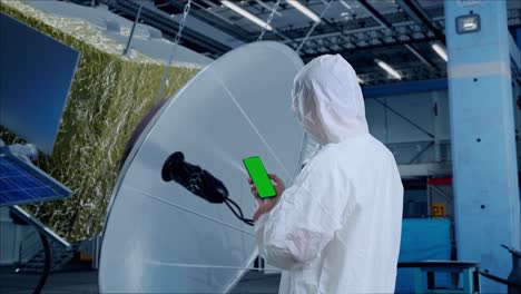 vista trasera de científico masculino asiático uso simulacro de pantalla verde teléfono inteligente mientras está de pie con la construcción de la nave espacial