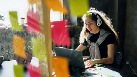 attractive female student is listening to music with headphones dancing and singing while working with laptop computer. glassboard with bright colored stickers in foreground.