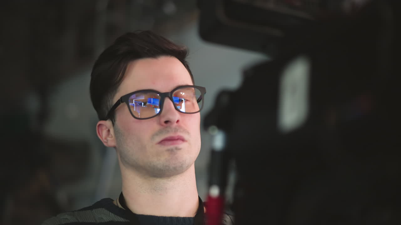 Close-up of man with glasses focused on professional camera in broadcast studio. Camera screen reflected in glasses. Operator wearing striped sweater, engaged in filming process