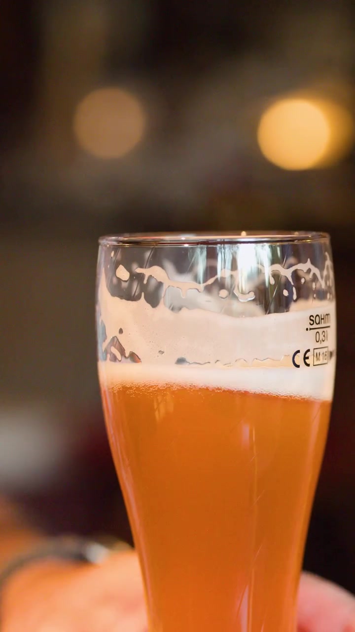 Una mano solleva una pinta di birra artigianale ambrata in un brewpub di Berlino illuminato calorosamente, profondità di campo ridotta.