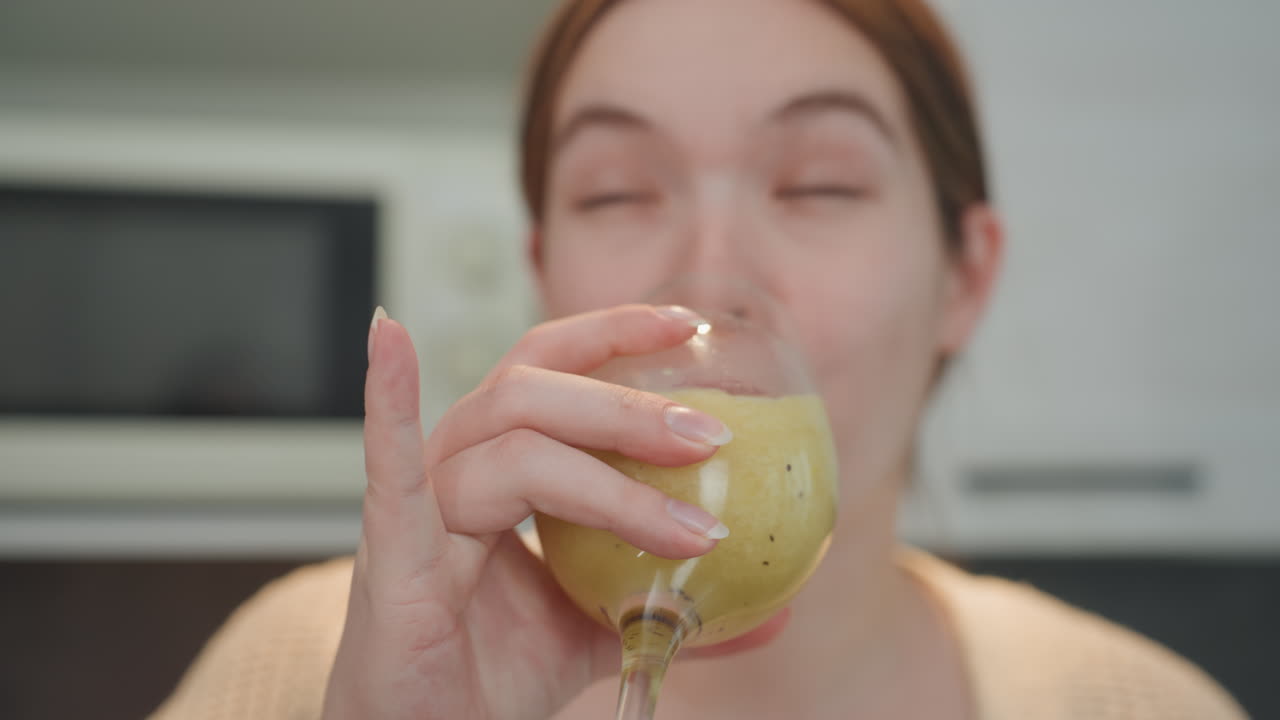 Mujer caucásica embarazada bebiendo un batido verde en una cocina moderna, primer plano de la cara y el vaso, saboreando la textura con una sonrisa sutil, cárdigan y bralette relajados, rutina de bienestar matutina enfocada.