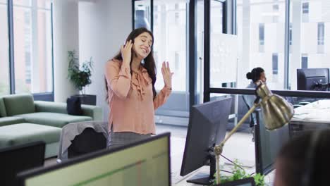 Smiling-caucasian-businesswoman-standing-and-talking-using-phone-headset-and-computer-in-busy-office